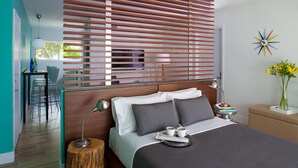 A modern bedroom with a wooden slat headboard, a bed with dark bedding, a bedside table with a lamp, and a small side table with a vase of flowers. - 1818 Meridian House Apartments and Suites by Eskape Collection (Miami Beach)