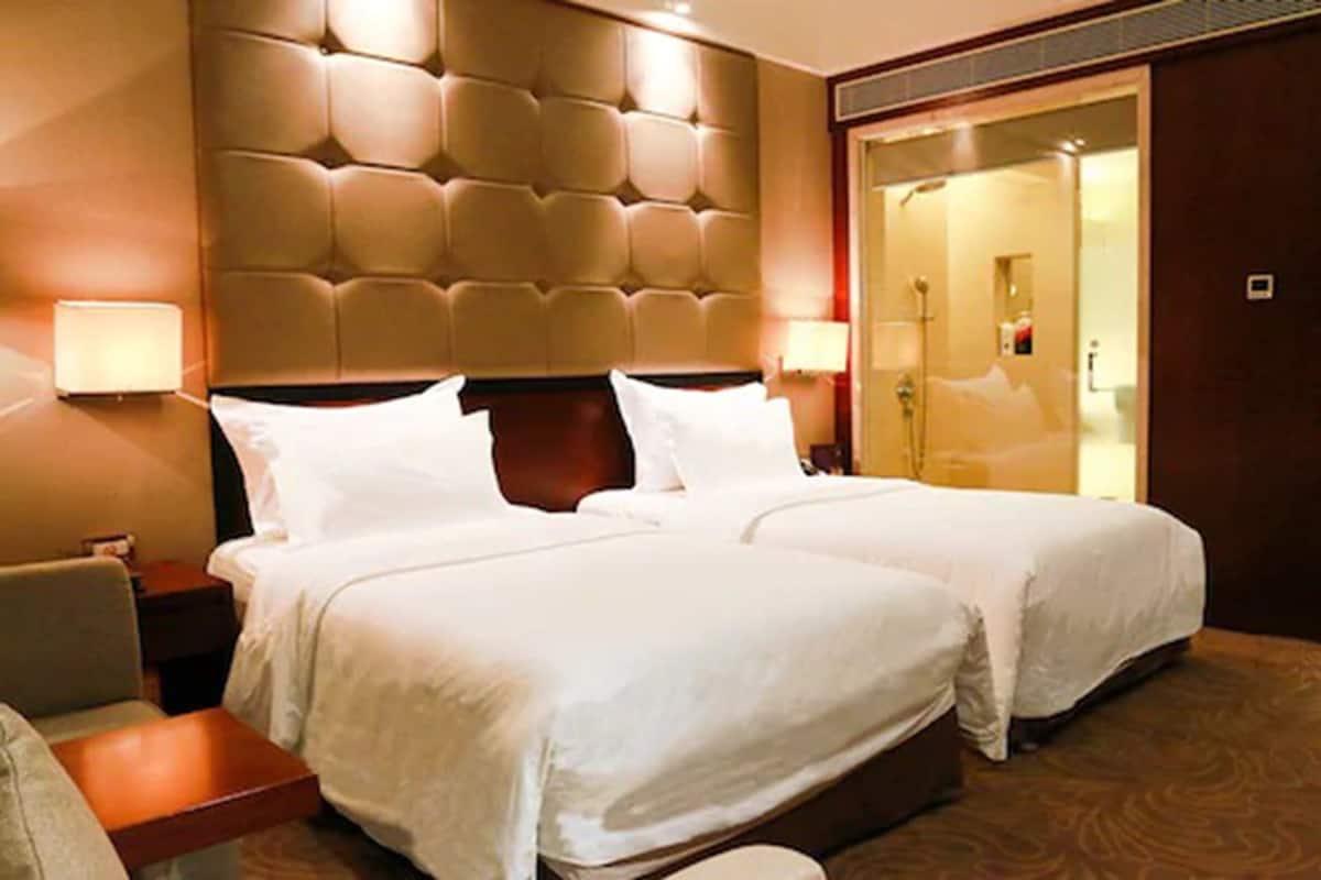 A hotel room with two beds, a padded headboard, bedside lamps, a nightstand, and a wooden bench.