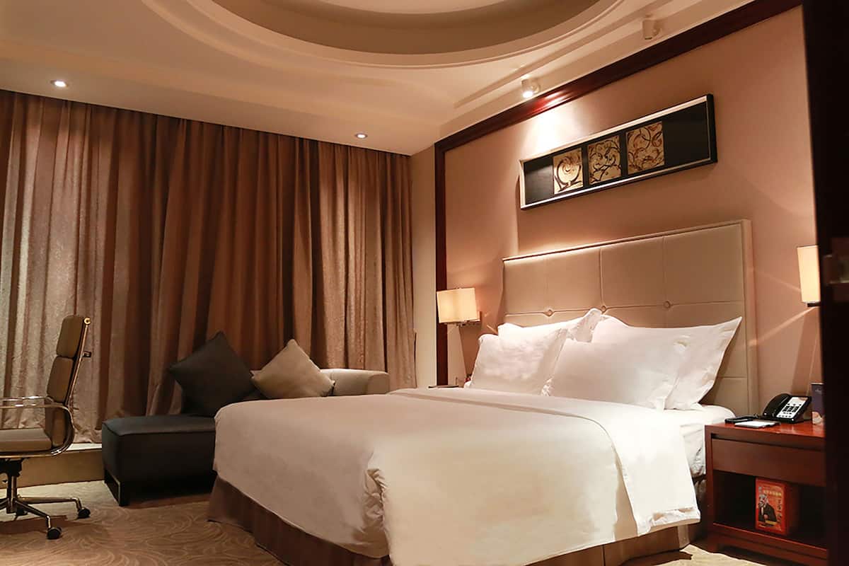 A hotel room with a large bed, a desk with a chair, a nightstand with a phone, and a wall-mounted artwork.