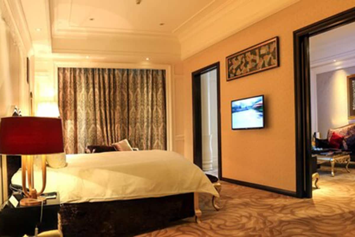 A hotel room with a bed, a TV, a desk with a lamp, and a view of another room.