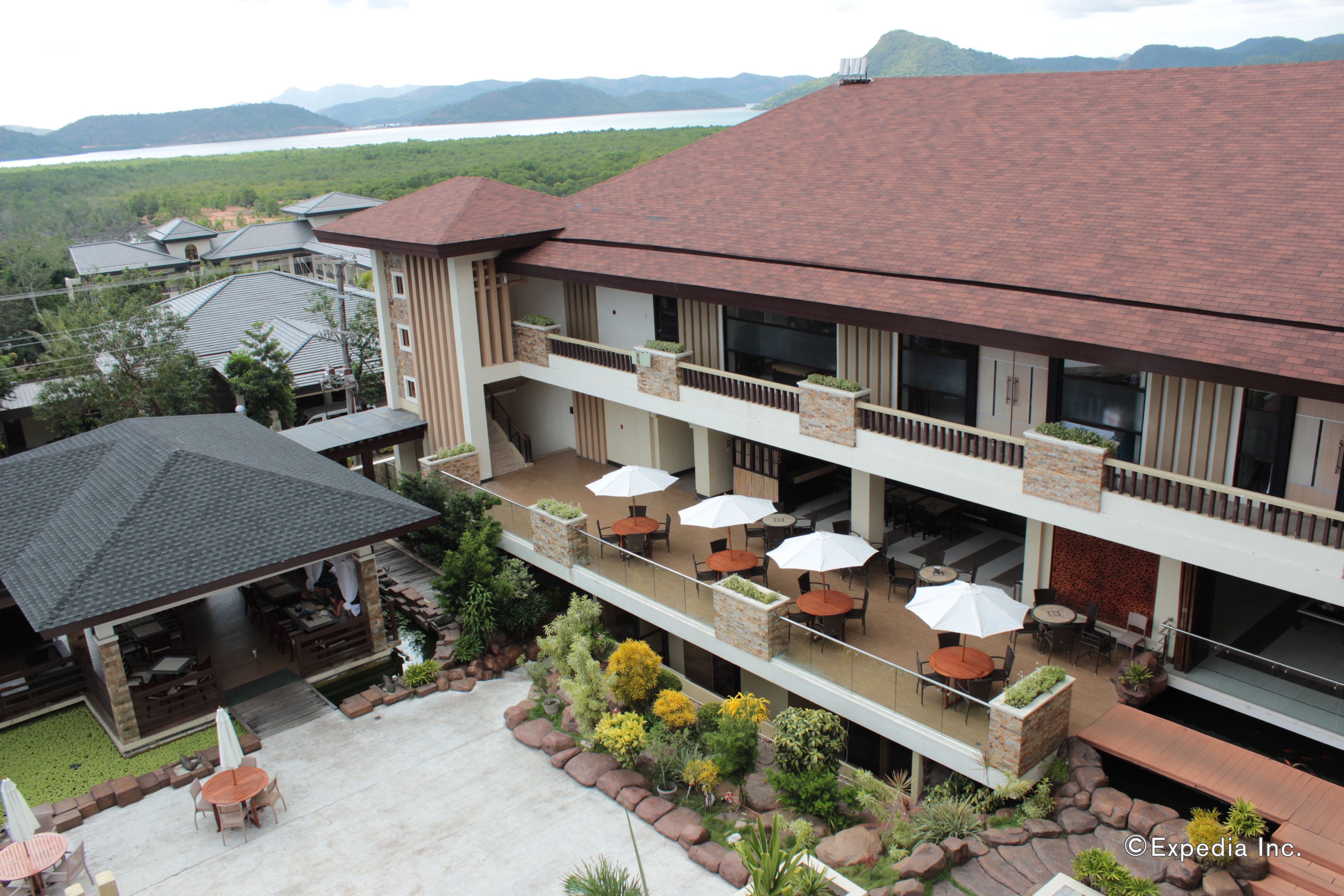 A hotel with a covered outdoor area, multiple dining tables with umbrellas, and a view of a lake and mountains.