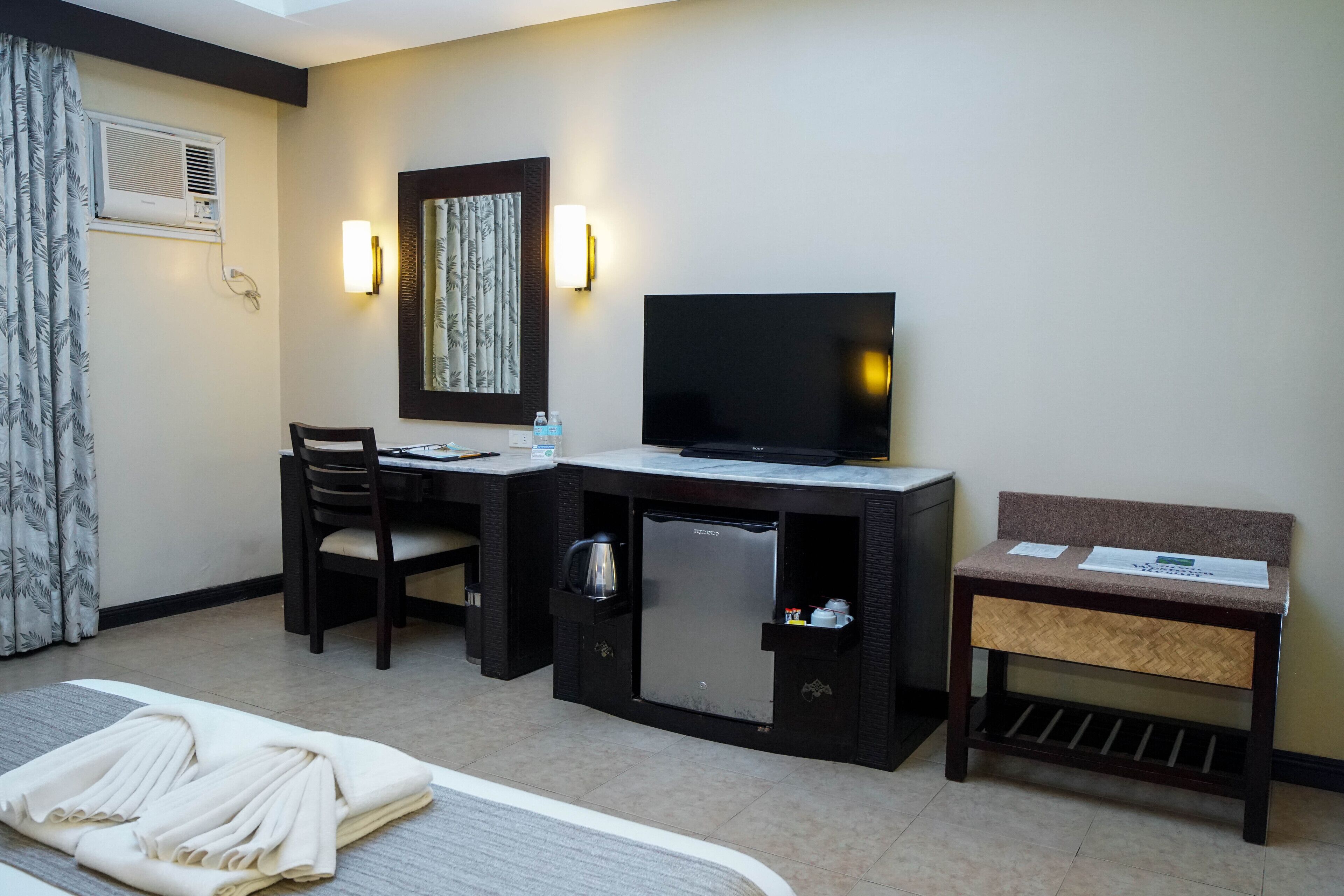 A hotel room with a bed, a desk with a television, a small refrigerator, a chair, and a bench.