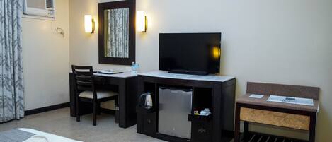 A hotel room with a bed, a desk with a television, a small refrigerator, a chair, and a bench.