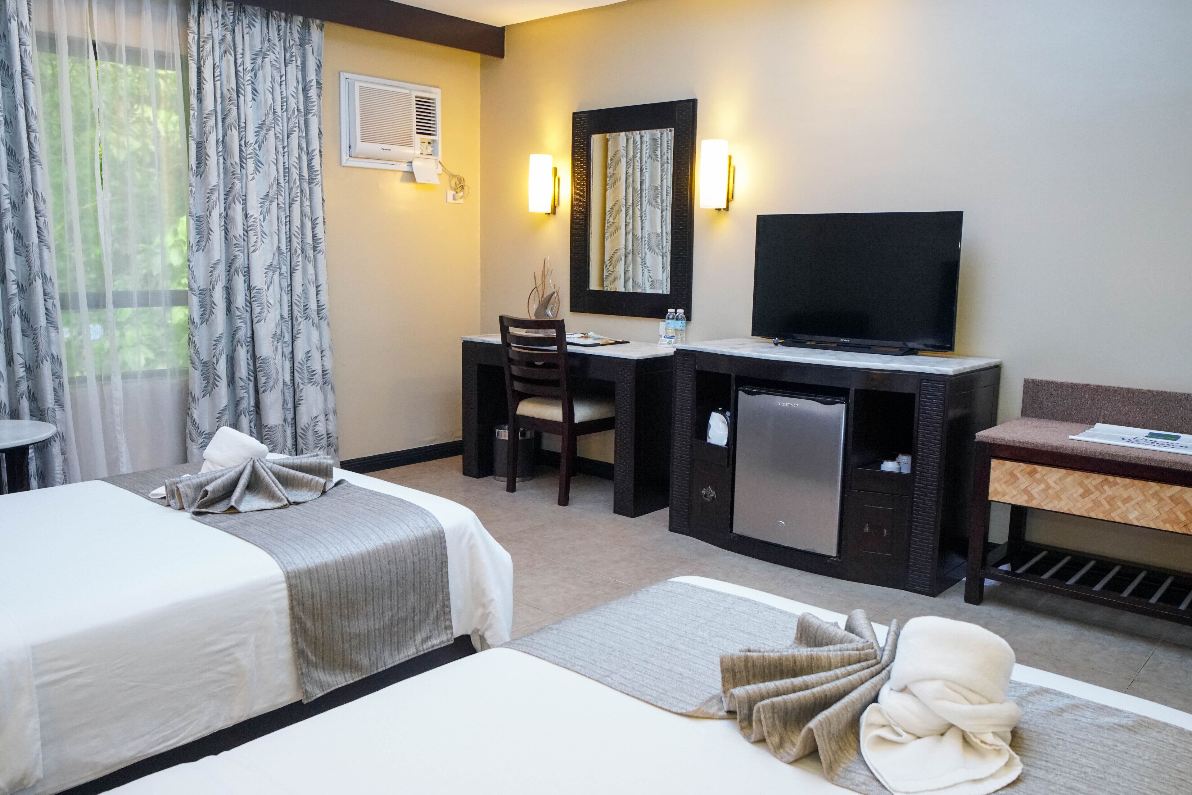 A hotel room with two beds, a desk with a television, a small refrigerator, and a seating area.