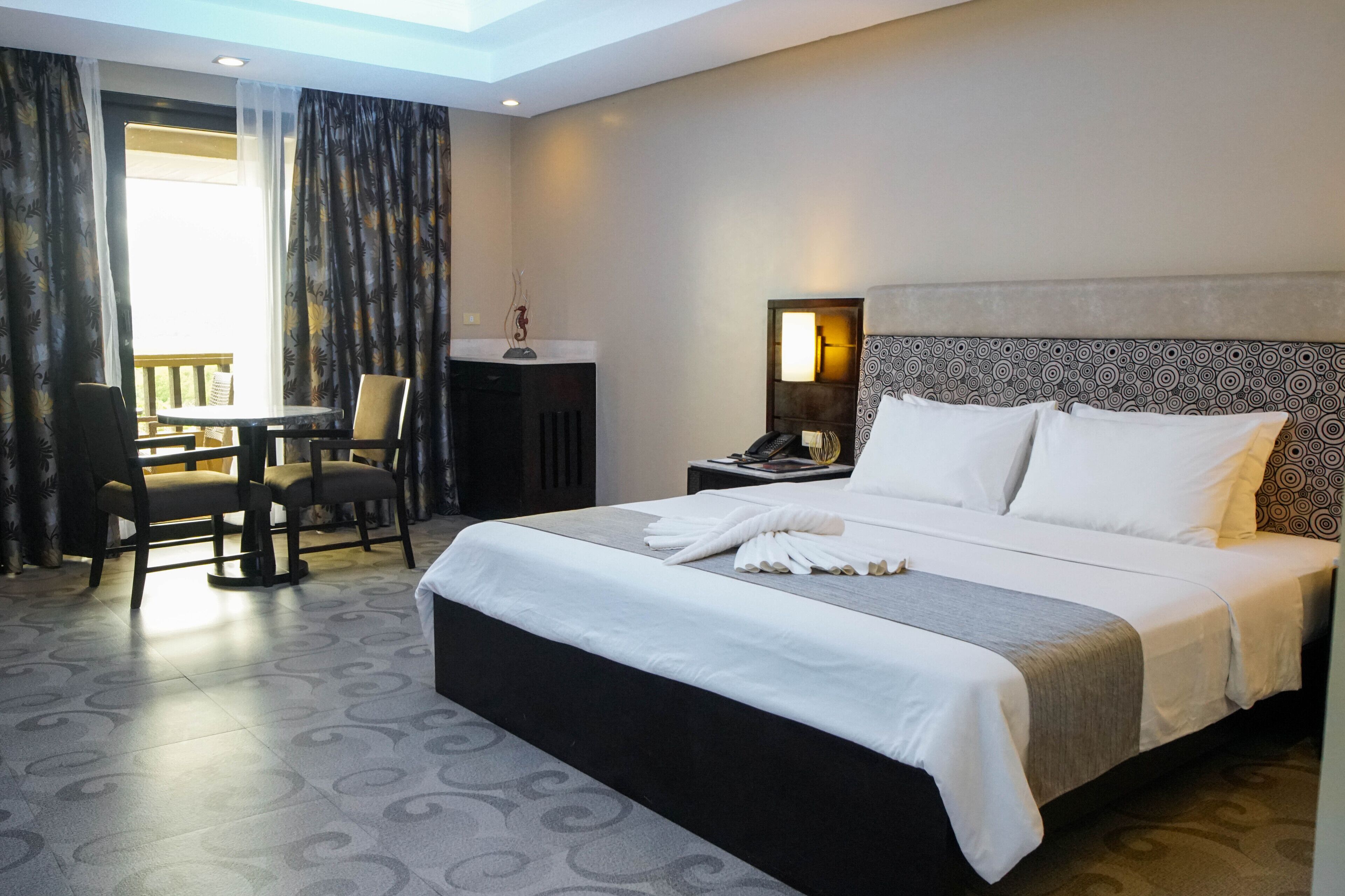 Une chambre d’hôtel avec un grand lit, un bureau avec une chaise, une petite table et un balcon avec des rideaux.