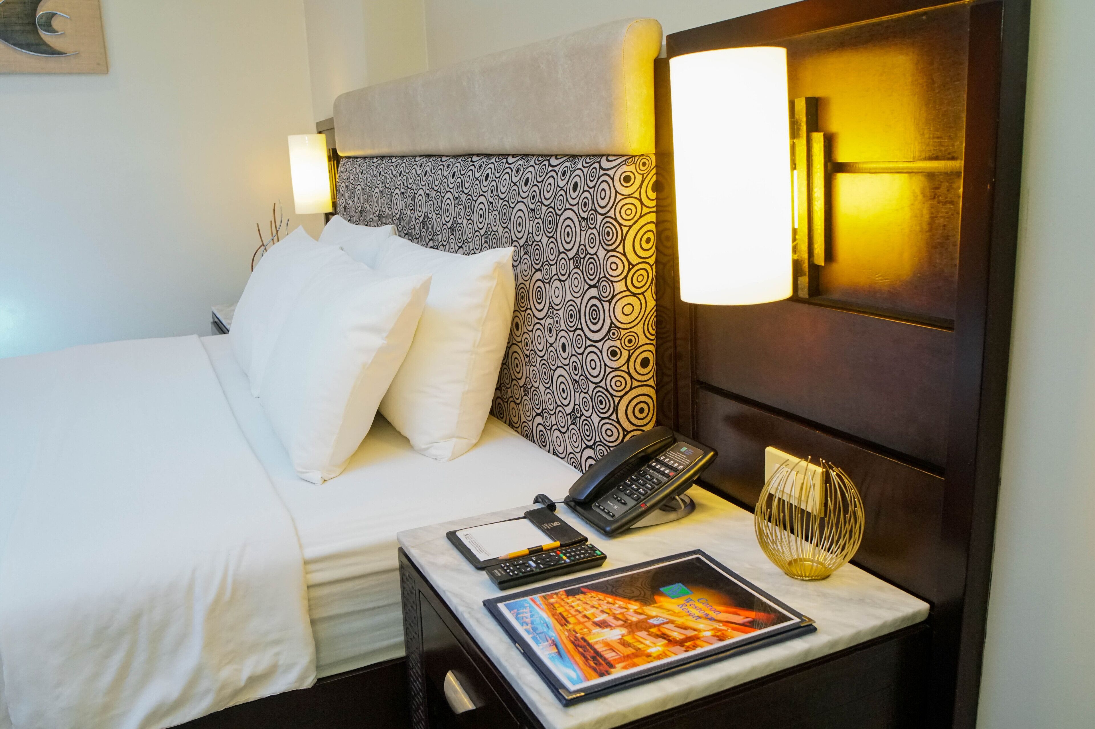 A hotel room with a bed, bedside table, lamp, phone, and a decorative item.