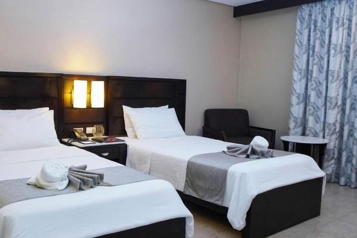 Habitación de hotel con dos camas, una mesita de noche con lámpara, una silla y una pequeña mesa.