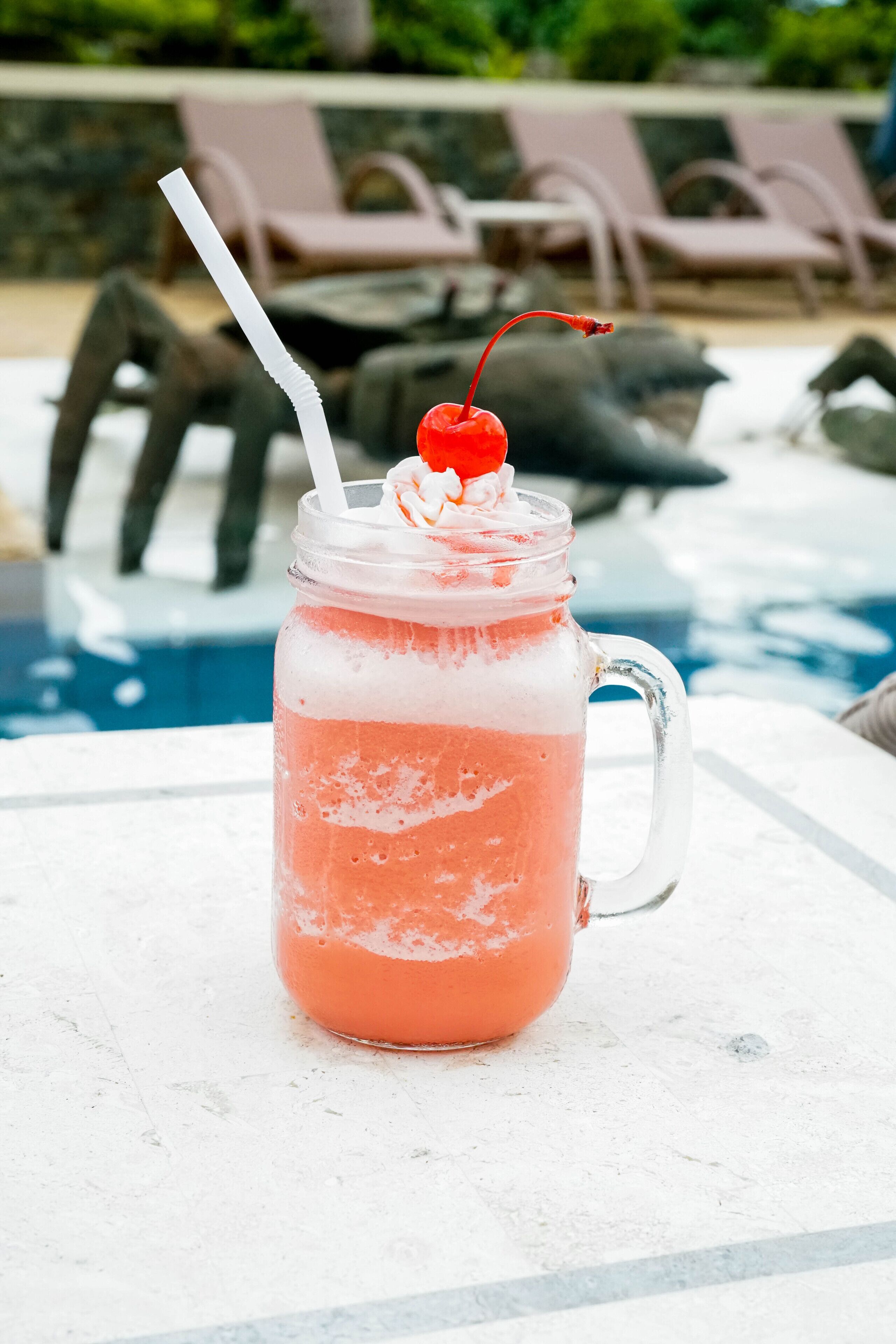 A pink drink in a glass mug with a straw and a cherry on top, placed on a white surface near a poolside area with lounge chairs.