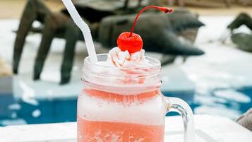 A pink drink in a glass mug with a straw and a cherry on top, placed on a white surface near a poolside area with lounge chairs.
