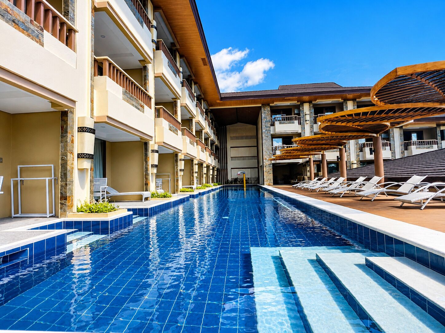 A swimming pool with steps leading into it, surrounded by lounge chairs and a building with balconies.