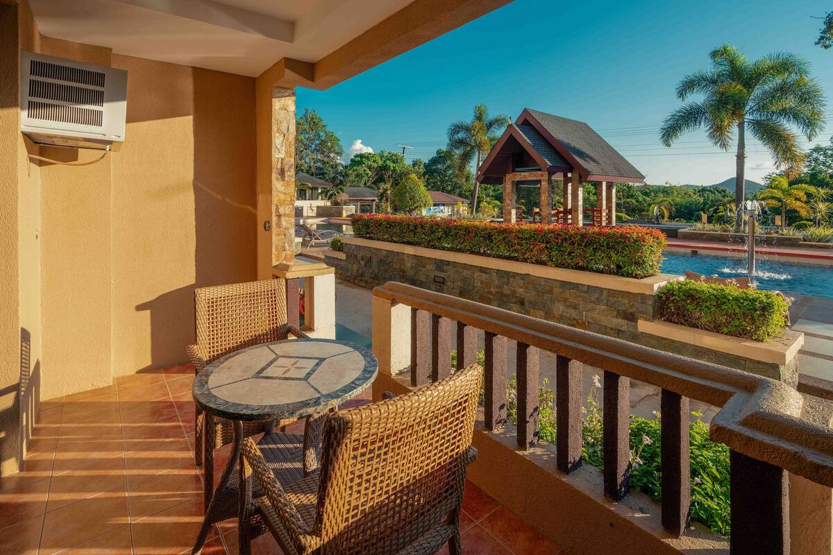 A balcony with wicker furniture, a stone wall, and a view of a pool and palm trees.