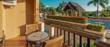 A balcony with wicker furniture, a stone wall, and a view of a pool and palm trees.