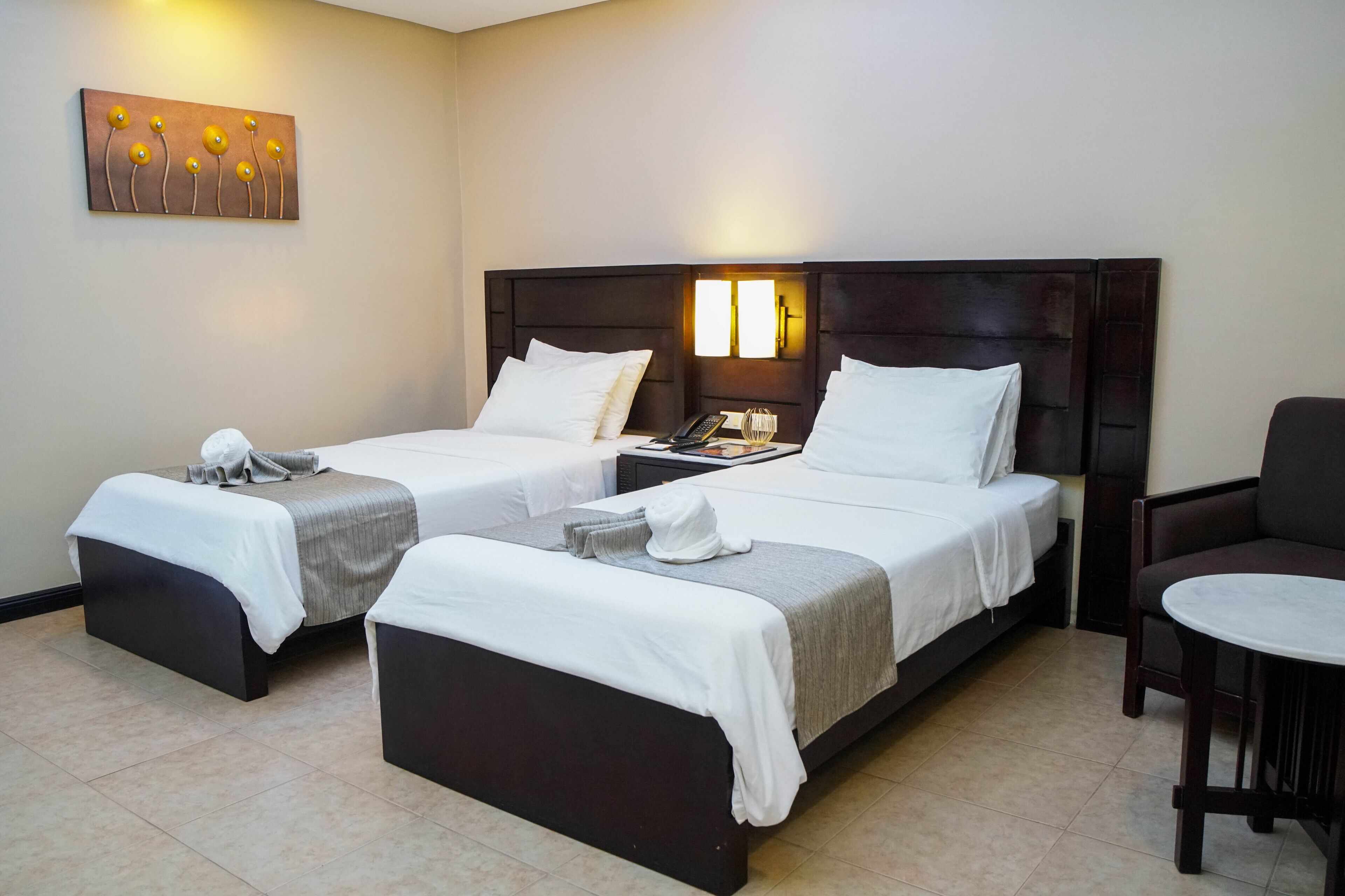 A hotel room with two single beds, a small table, a chair, and a wall-mounted artwork.
