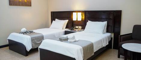 A hotel room with two single beds, a small table, a chair, and a wall-mounted artwork.