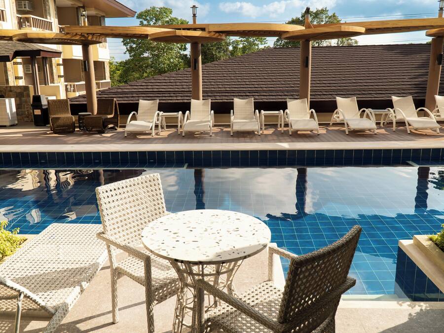 A pool area with lounge chairs and a table, surrounded by a building with a tiled roof and balconies.