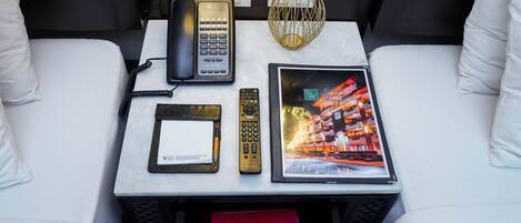 A hotel room with a white bed, a bedside table with a phone, remote controls, a framed picture, and a red notebook.