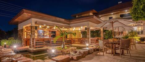 An outdoor patio area with stone seating, tables, and umbrellas, surrounded by a pool and a building with a covered seating area.