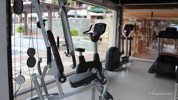 Un gimnasio con diversos equipos de ejercicio, una ventana con vista a una piscina y edificios, y un ventilador de techo.