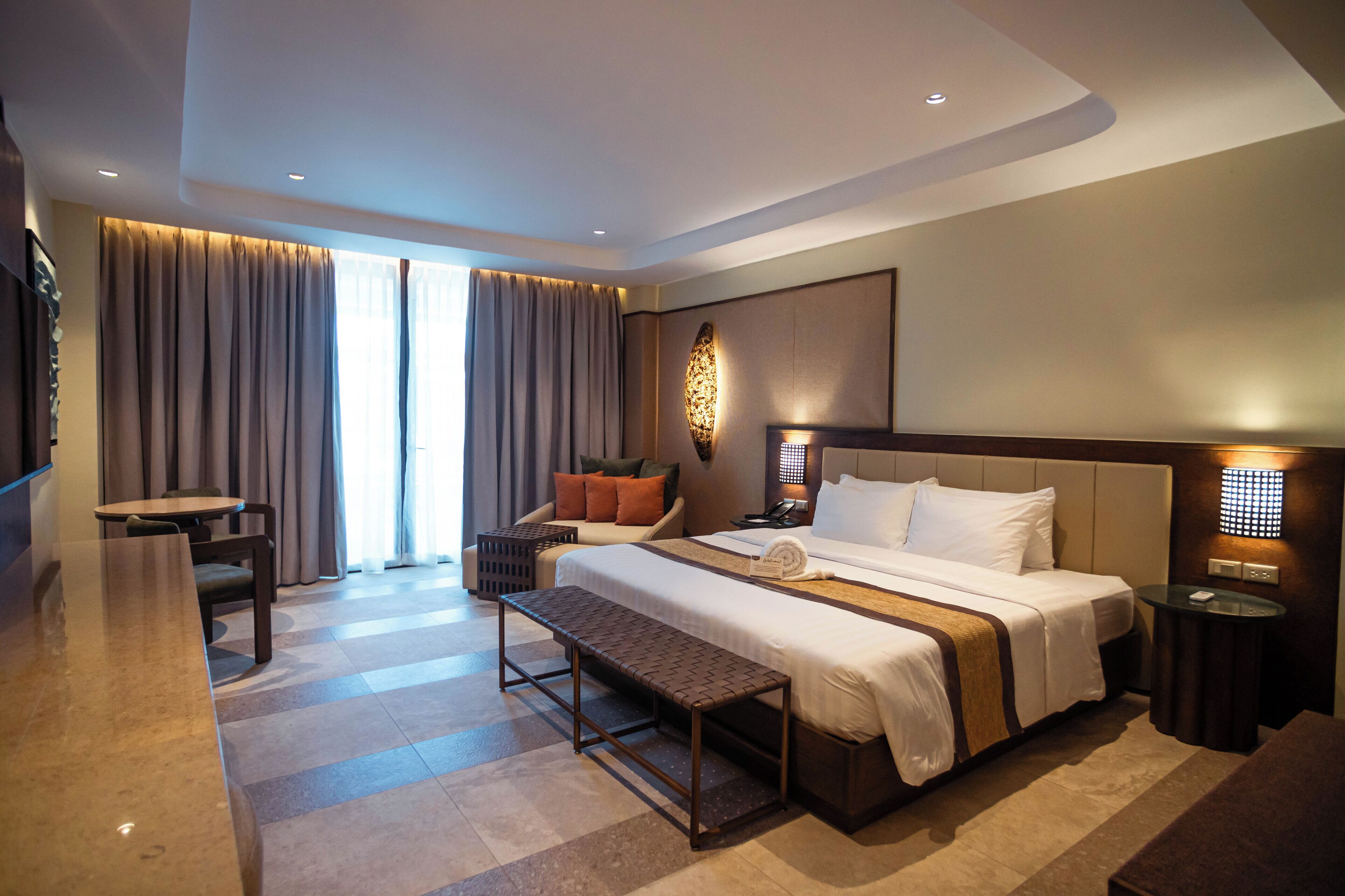 A hotel room with a large bed, a desk, a chair, a bench, a bedside table, a lamp, and a wall-mounted light fixture.