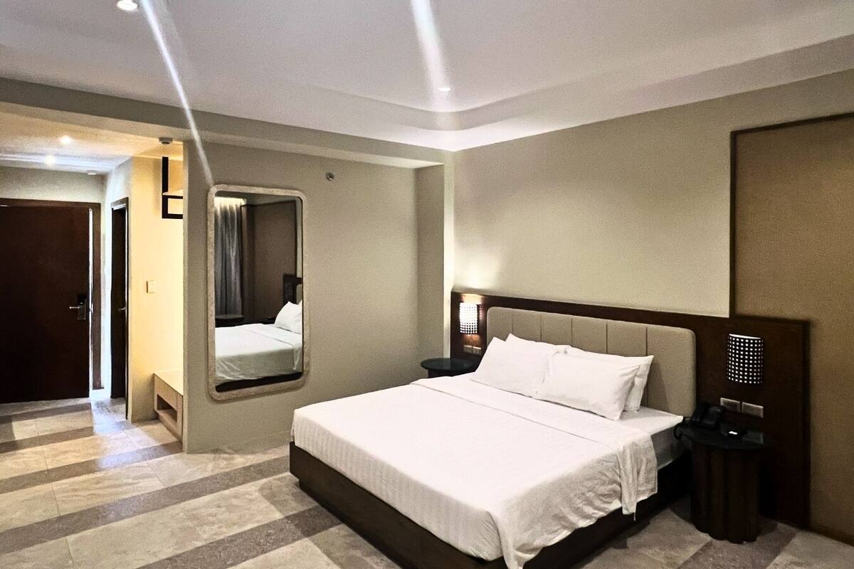 A hotel room with a large bed, a bedside table with a lamp, a mirror, and a door leading to another room.