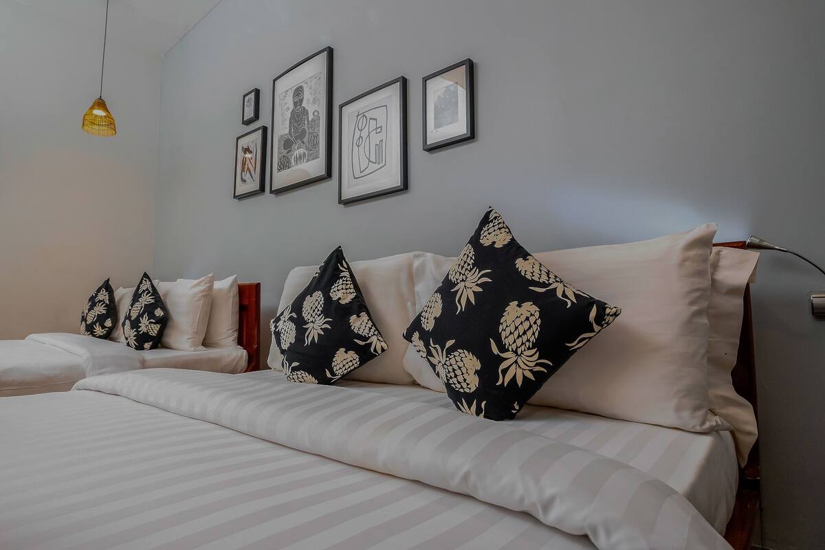 Habitación de hotel con dos camas, cada una con ropa de cama blanca y almohadas estampadas en blanco y negro. En la pared hay obras de arte enmarcadas y una lámpara colgante.