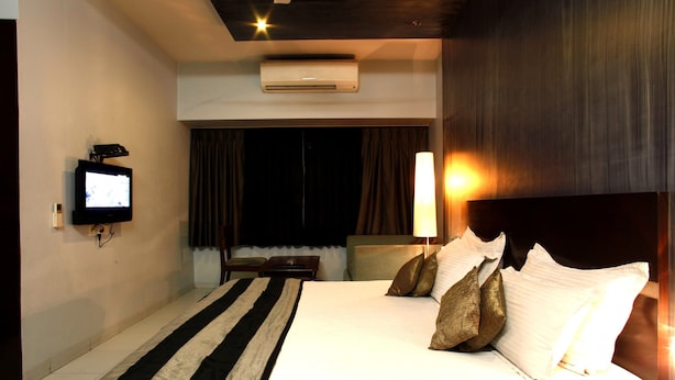 A hotel room with a bed, a TV mounted on the wall, a bedside table, a lamp, and an air conditioning unit. - Acacia Inn (Jaipur)
