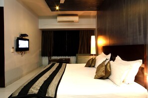 A hotel room with a bed, a TV mounted on the wall, a bedside table, a lamp, and an air conditioning unit. - Acacia Inn (Jaipur)
