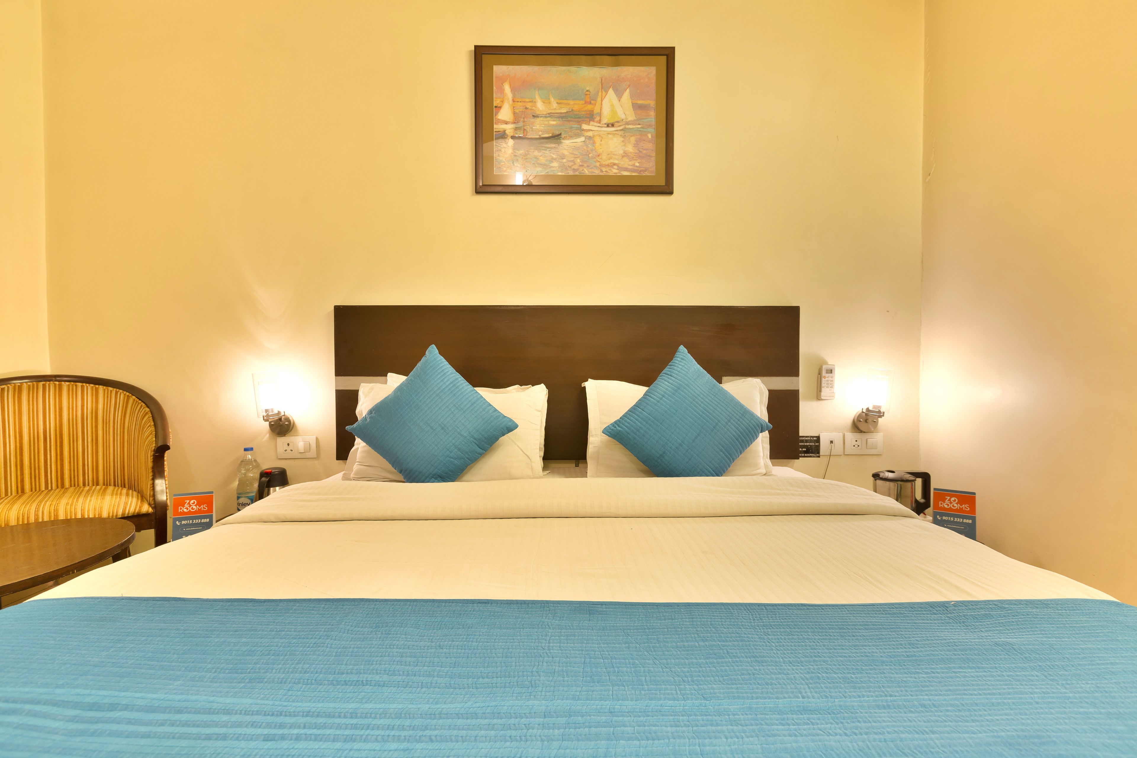 A hotel room with a large bed, two blue pillows, a headboard, a painting on the wall, a chair, and a nightstand with a lamp.