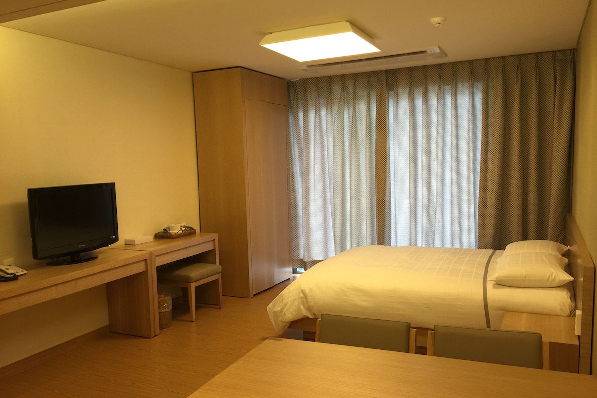 Una habitación de hotel con cama, escritorio con televisor, silla y ventana con cortinas.