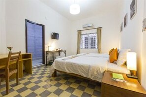 A hotel room with a checkered floor, a bed with pillows, a desk with a chair, a television, and a window with curtains.