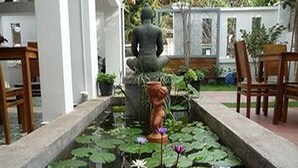 Un patio tranquilo con una estatua, un pequeño estanque con nenúfares y flores, y sillas de madera.