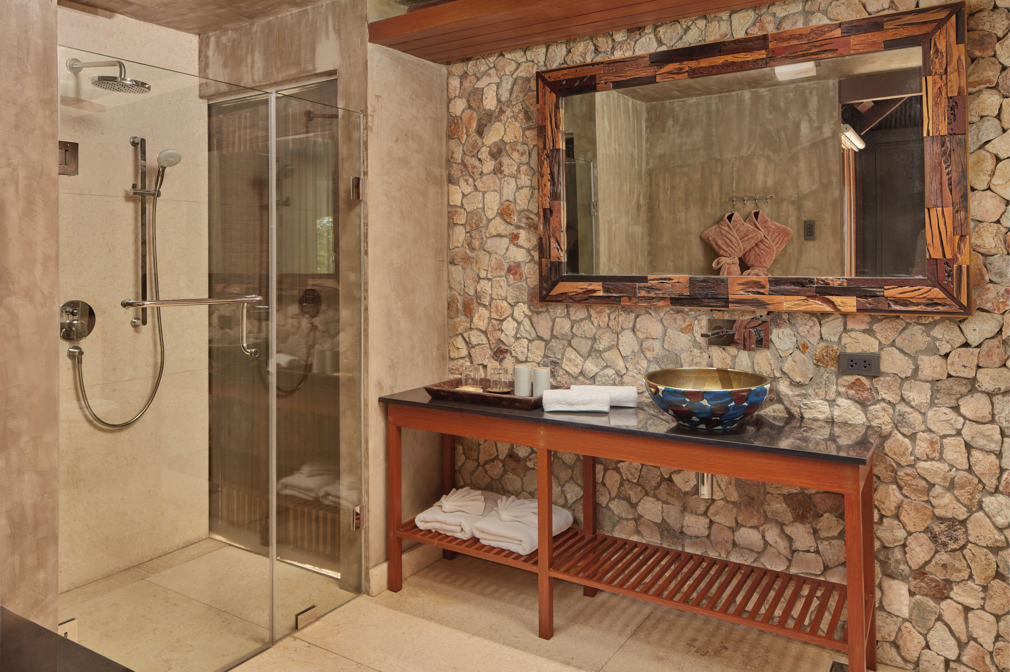 A bathroom with a glass-enclosed shower, a stone wall, a wooden vanity, and a mirror with a wooden frame.