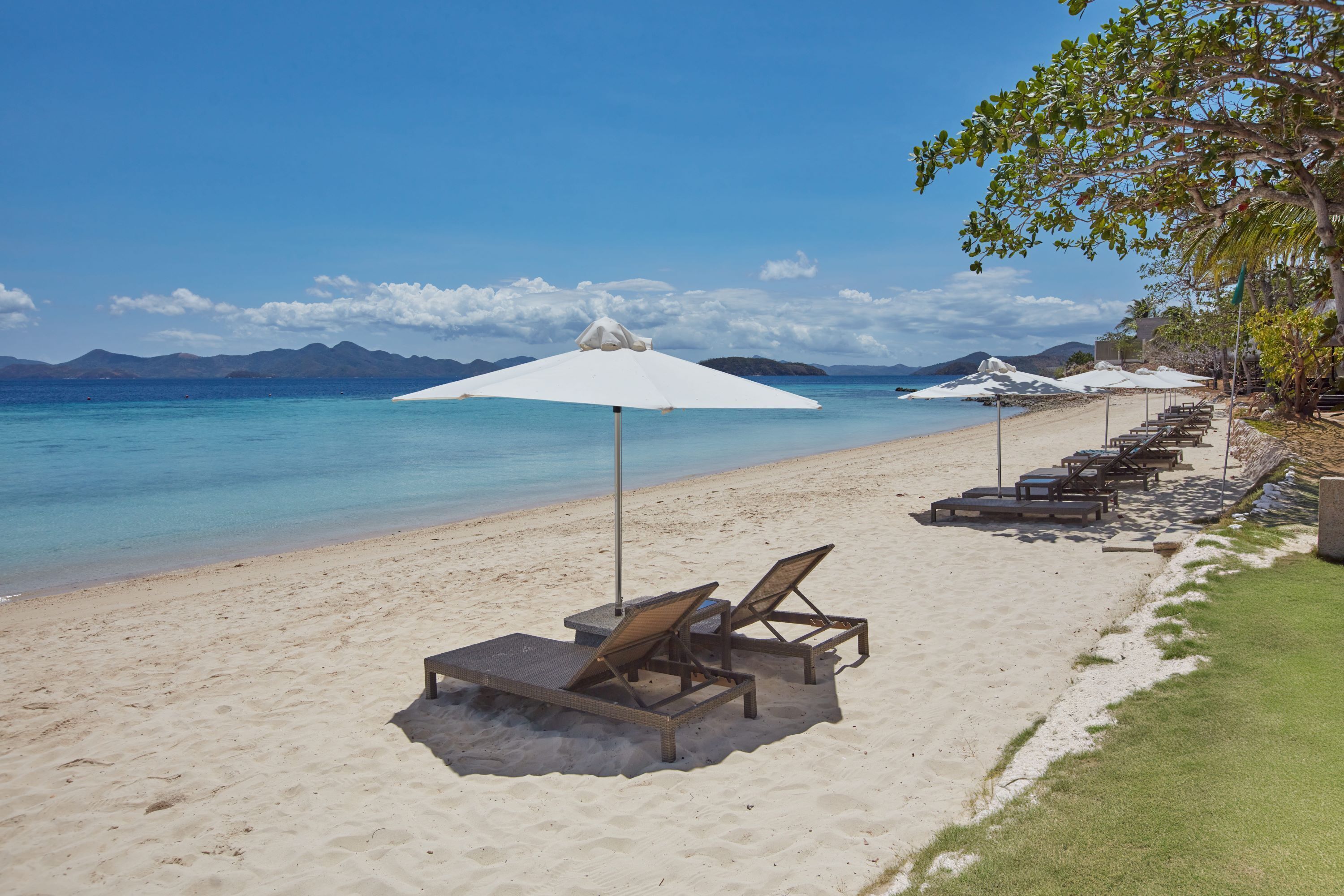 A beach with white umbrellas and lounge chairs, a clear blue sky, and mountains in the background.