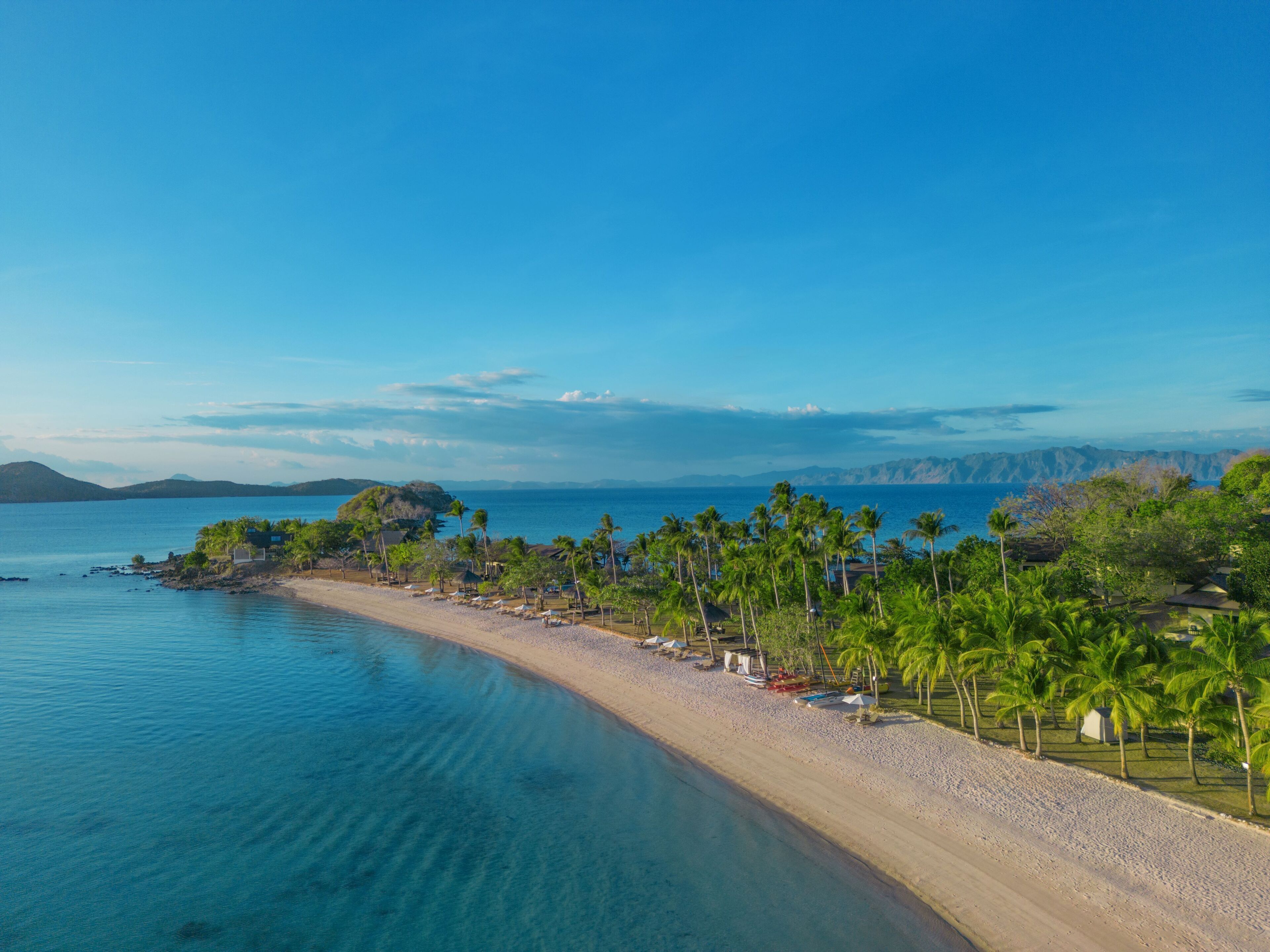A beach with clear blue water, palm trees, and a view of distant mountains.