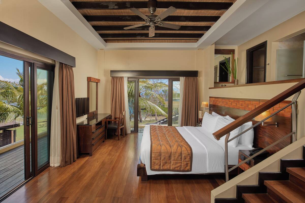 A spacious bedroom with a large bed, wooden flooring, a ceiling fan, and a balcony with curtains.