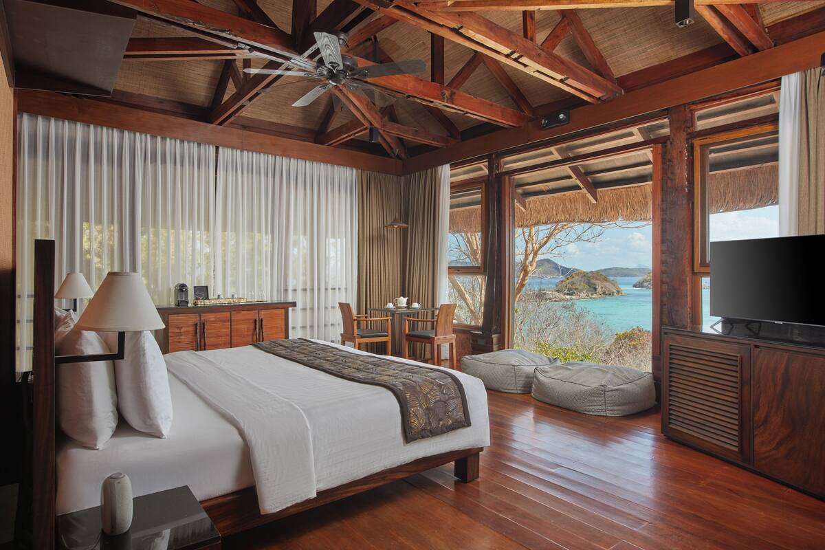 A spacious bedroom with a large bed, wooden ceiling beams, a television, and a view of the ocean.