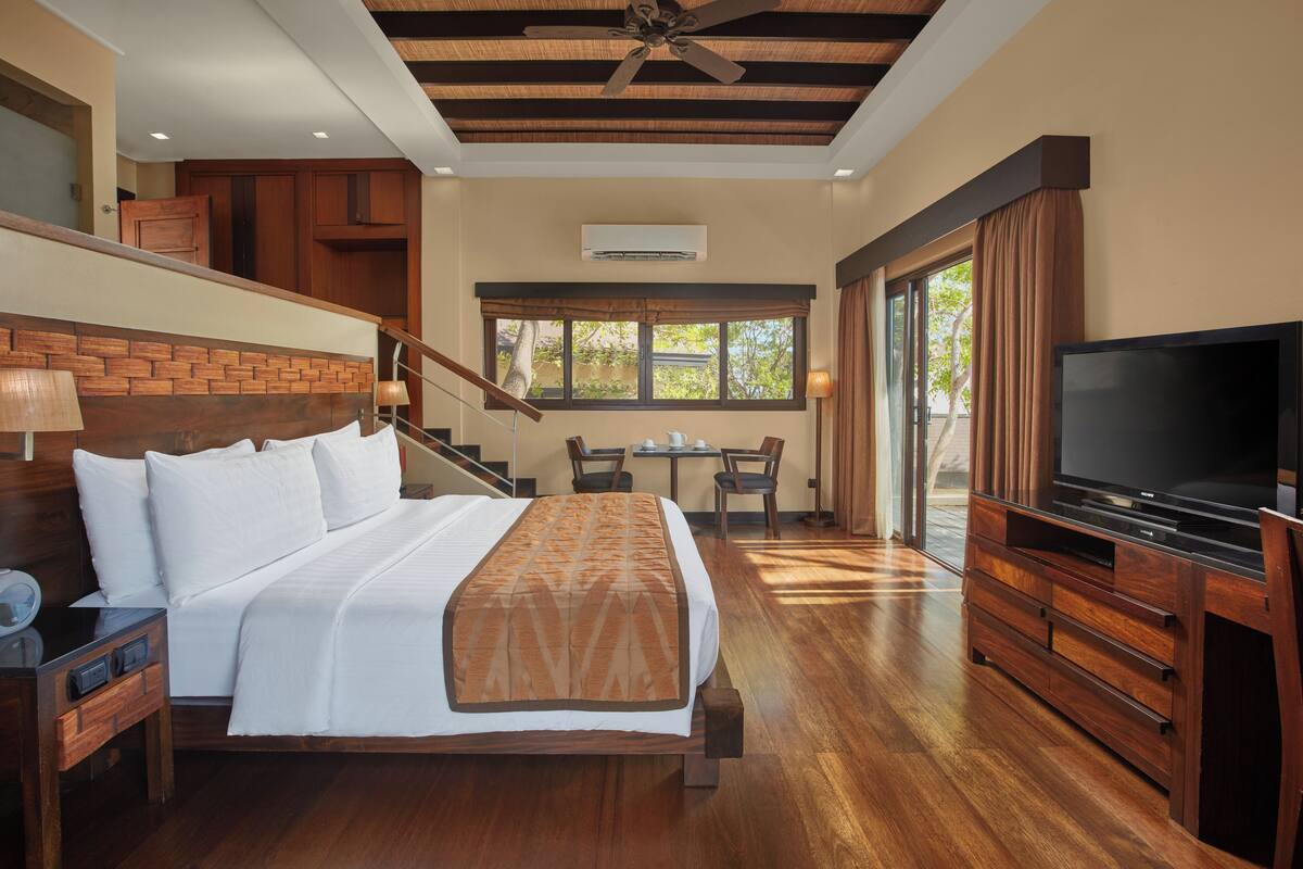 A hotel room with a large bed, wooden headboard, a TV, a desk with chairs, and a view of the outdoors.