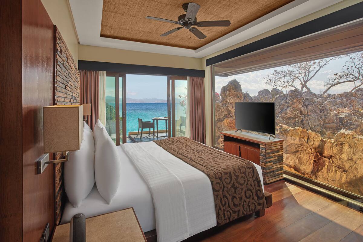 A hotel room with a large bed, a TV, a balcony with a view of the sea, and a stone wall.