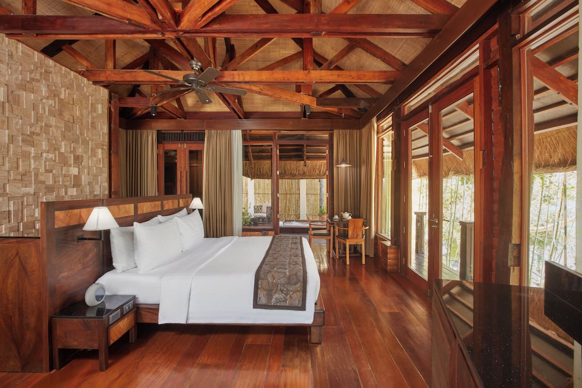 A spacious bedroom with a large bed, wooden ceiling beams, a ceiling fan, and a view through large windows.