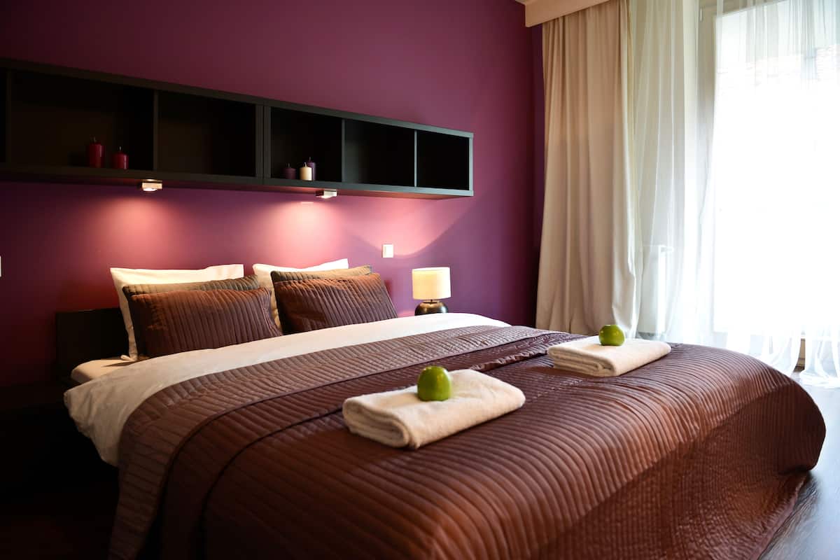 A hotel room with a large bed, a bedside table with a lamp, a shelf above the bed, and a window with curtains.