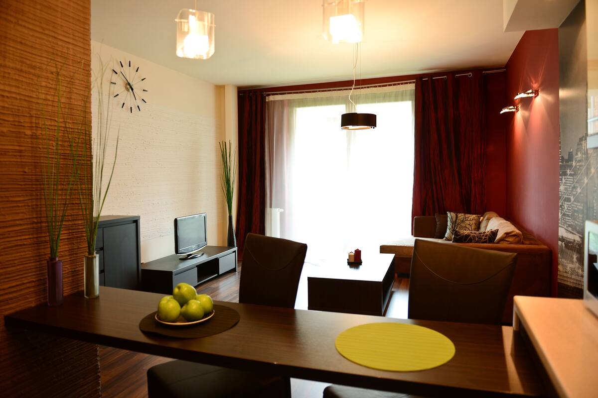 A modern living room with a dining area, featuring a television, seating, and a large window with curtains.