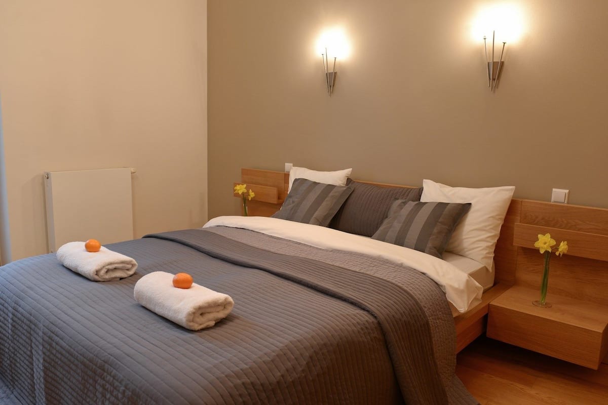 A neatly made bed with a gray and white striped bedspread, two pillows, and two folded towels with oranges. There are two wall-mounted light fixtures on either side of the bed.