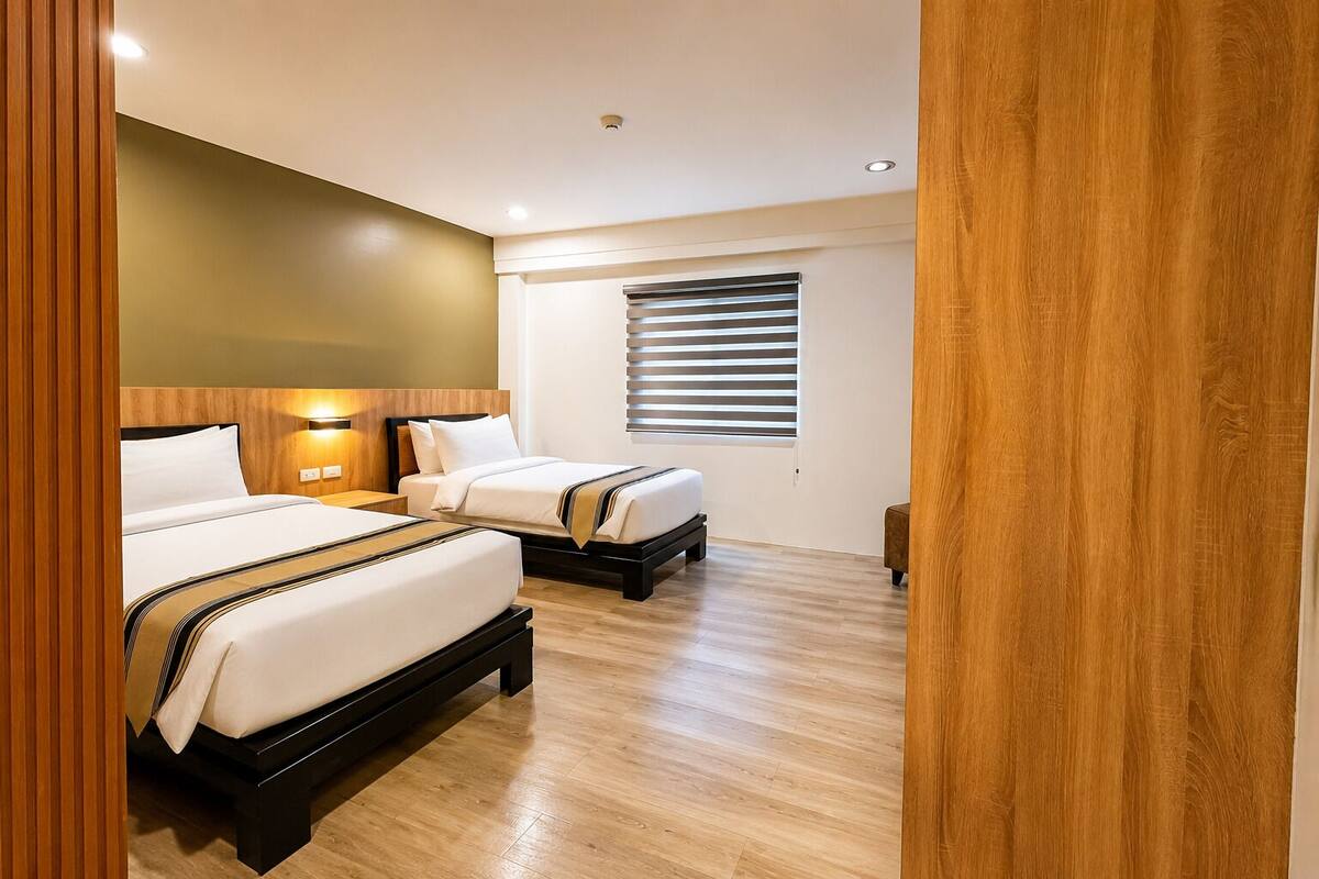 A hotel room with two beds, wooden flooring, a window with blinds, and a wall-mounted light fixture.