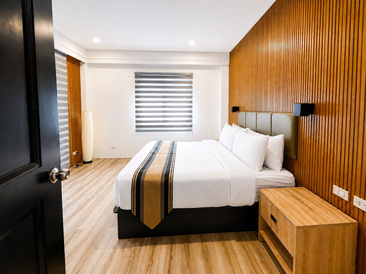 A modern hotel room with a bed, wooden panel wall, and a window with blinds.