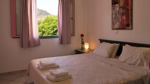 A bedroom with a bed, pillows, a nightstand with a lamp, and a window with curtains. - Cybele Suites & Apartments (Ierapetra)