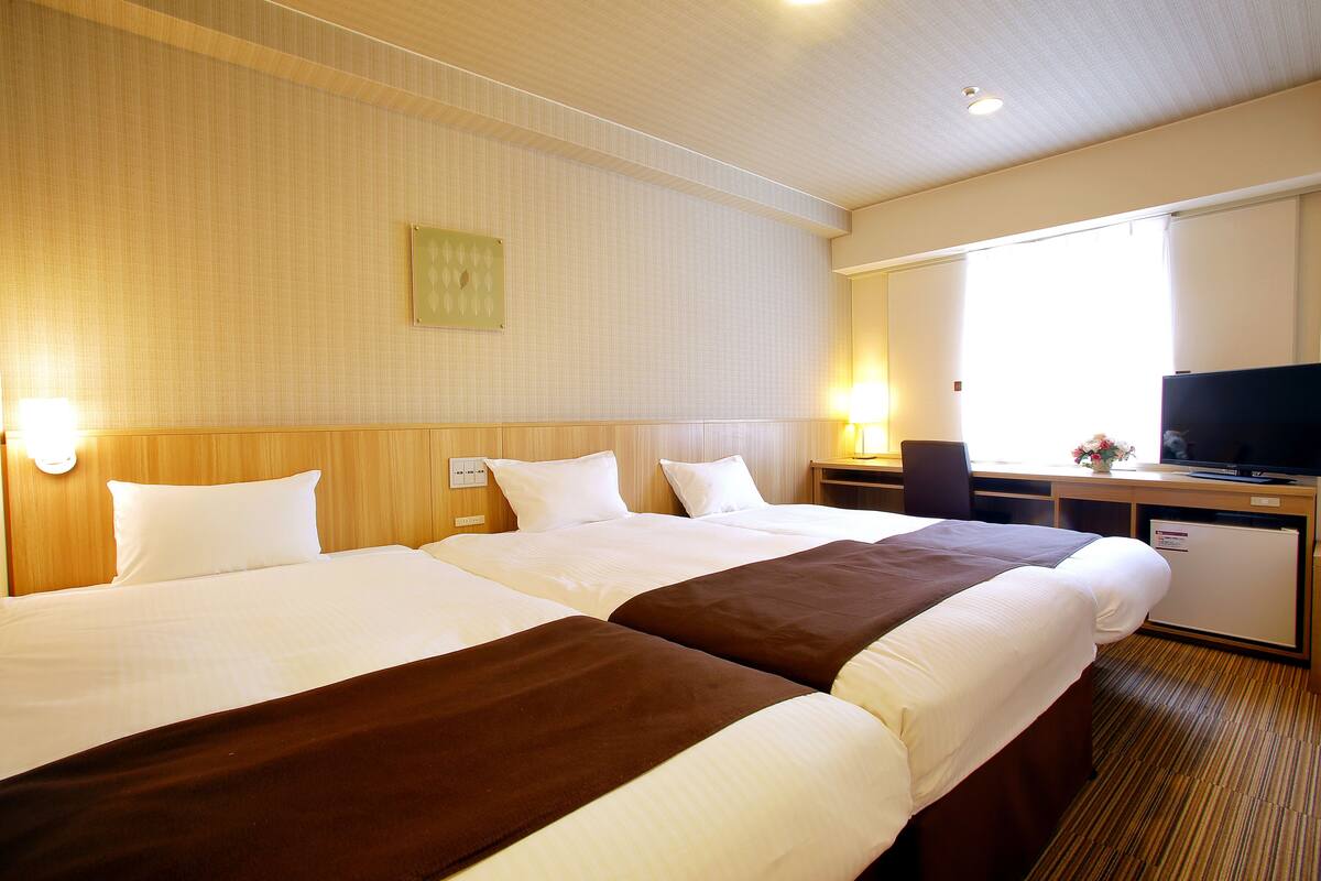 Ein Hotelzimmer mit zwei Betten, einem Schreibtisch, einem Fernseher und einem Fenster mit Vorhängen.
