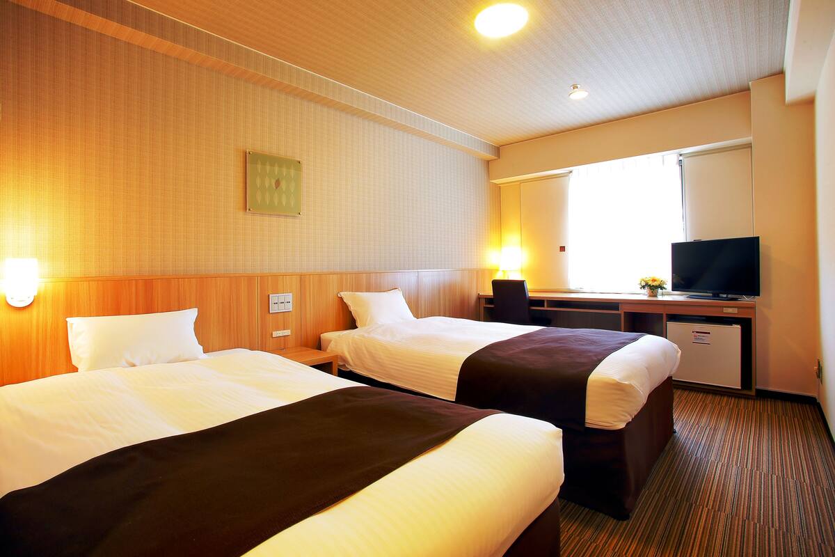 Ein Hotelzimmer mit zwei Betten, einem Schreibtisch, einem Fernseher und einem kleinen Kühlschrank.