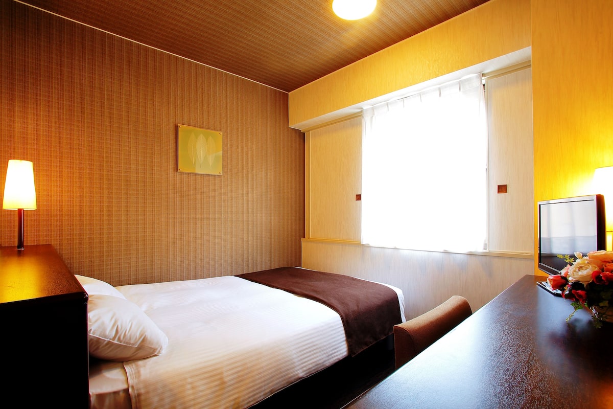 Ein Hotelzimmer mit zwei Betten, einem Holz-Kopfteil, einem Nachttisch mit Lampe, einem Fenster mit Vorhängen und einem kleinen Bild an der Wand.