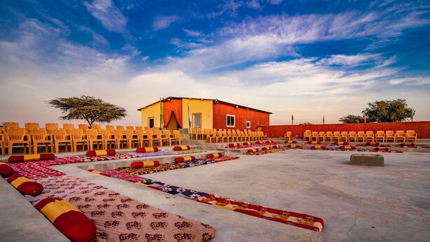 Un patio con filas de sillas y alfombras coloridas, un edificio con fachada roja y un cielo despejado.