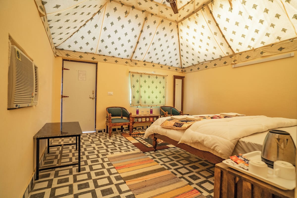 Una habitación con piso estampado, una cama con edredón blanco, un escritorio con dos sillas, una ventana con cortinas verdes y un ventilador de techo.